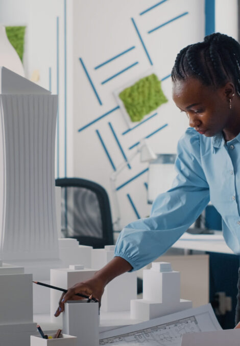 African american woman analyzing blueprints plan and building model to work on architecture African american woman analyzing blueprints plan and building model to work on architecture. Architect working with maquette and layout print to design urban construction for development.