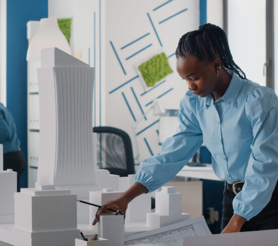 African american woman analyzing blueprints plan and building model to work on architecture. Architect working with maquette and layout print to design urban construction for development.