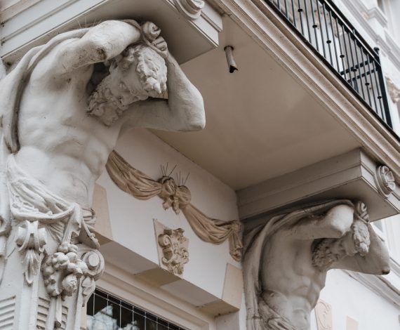 Two stone male sculptures with muscular bodies hold up a balcony on the facade of a historic European-style building